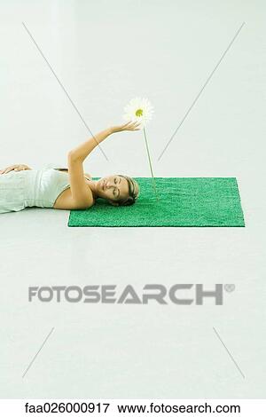Stock Photo - Woman lying on artificial turf, touching gerbera daisy, eyes closed. Fotosearch
