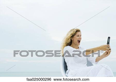 Woman photographing self with cell phone outdoors, open mouth View Large Photo Image Stock Photograph - Woman photographing self with cell phone outdoors, open mouth. Fotosearch