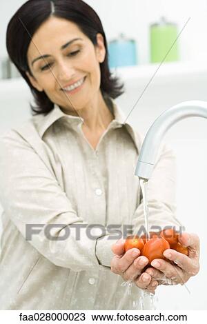 Stock Image - Woman rising handful of tomatoes under faucet, smiling. Fotosearch