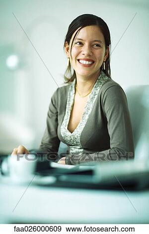 Stock Photo - Woman sitting at desk, smiling, looking away. Fotosearch