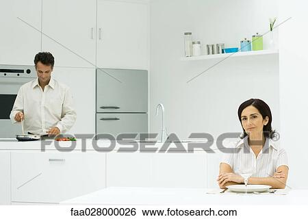 Woman sitting at table with arms folded, looking at camera, husband preparing meal in the background View Large Photo Image Stock Photograph - Woman sitting at table with arms folded, looking at camera, husband preparing meal in the background. Fotosearch
