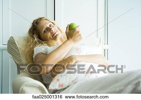 Woman sitting in armchair, holding apple, smiling at camera View Large Photo Image Stock Photograph - Woman sitting in armchair, holding apple, smiling at camera. Fotosearch