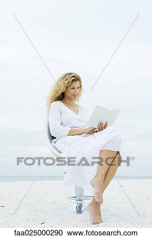 Stock Image - Woman sitting in chair at the beach, reading book. Fotosearch