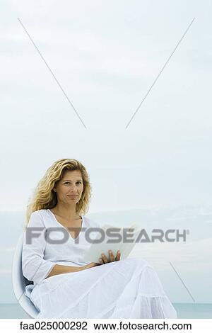 Woman sitting in chair at the beach, holding book, smiling at camera View Large Photo Image Stock Image - Woman sitting in chair at the beach, holding book, smiling at camera. Fotosearch