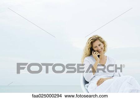 Woman sitting in chair outdoors, using cell phone, smiling View Large Photo Image Picture - Woman sitting in chair outdoors, using cell phone, smiling. Fotosearch