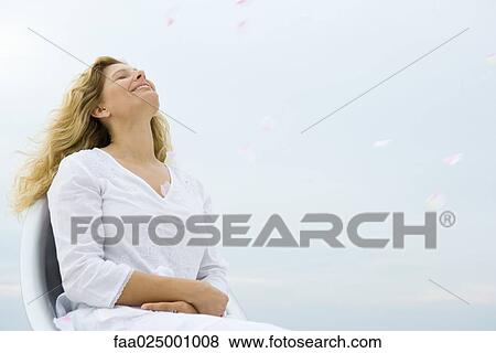 Stock Photo - Woman sitting with eyes closed and head back, smiling, sky in background, low angle view. Fotosearch