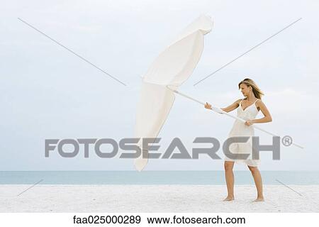 Stock Photo - Woman standing on beach, opening large beach umbrella. Fotosearch