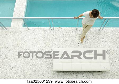 Stock Photo - Woman standing on terrace, leaning against railing, swimming pool in background. Fotosearch