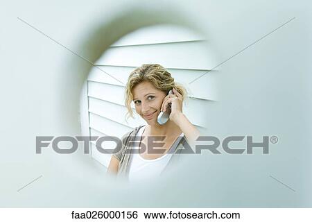 Woman using cell phone, smiling at camera, viewed through hole View Large Photo Image Stock Photograph - Woman using cell phone, smiling at camera, viewed through hole. Fotosearch