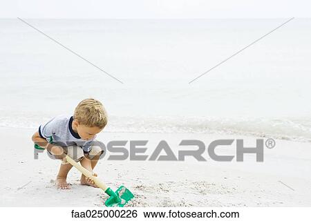 Young boy crouching at the beach, digging in sand with shovel View Large Photo Image Stock Photograph - Young boy crouching at the beach, digging in sand with shovel. Fotosearch