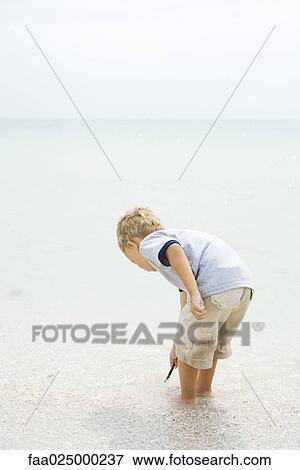 Young boy standing ankle deep in water at the beach, bending over, looking down View Large Photo Image Stock Photo - Young boy standing ankle deep in water at the beach, bending over, looking down. Fotosearch