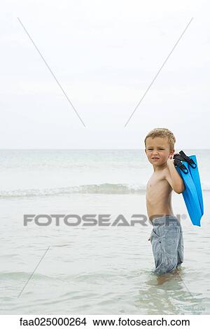 Young boy standing knee deep in water, carrying flippers, looking over shoulder at camera View Large Photo Image Picture - Young boy standing knee deep in water, carrying flippers, looking over shoulder at camera. Fotosearch