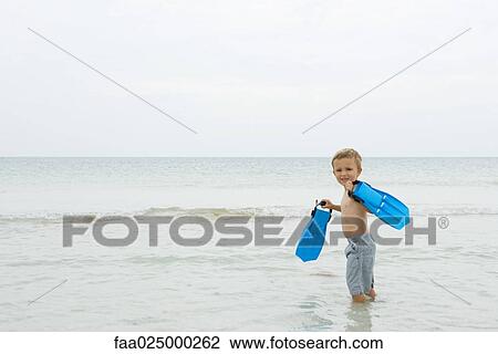 Stock Image - Young boy standing knee deep in water, carrying flippers, smiling over shoulder at camera. Fotosearch