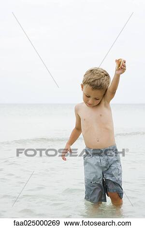 Young boy standing knee deep in water, holding up seashell, looking down View Large Photo Image Stock Photo - Young boy standing knee deep in water, holding up seashell, looking down. Fotosearch