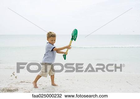 Stock Image - Young boy standing on beach, holding up shovel, looking down. Fotosearch