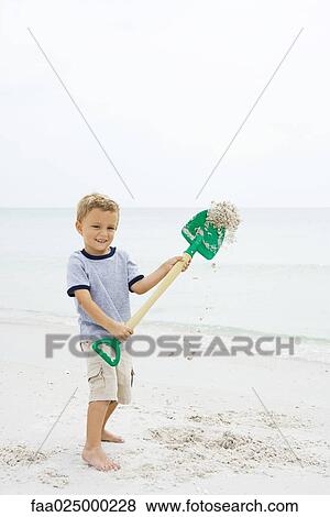 Stock Photo - Young boy standing on beach, holding up shovel, throwing sand. Fotosearch