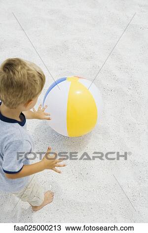 Stock Image - Young boy standing on the beach, reaching for beach ball, high angle view. Fotosearch