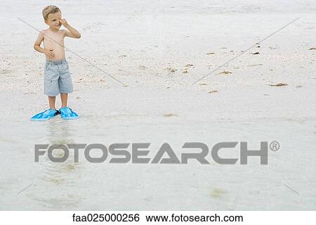 Stock Photograph - Young boy wearing flippers at the beach, standing at water's edge, rubbing eyes. Fotosearch