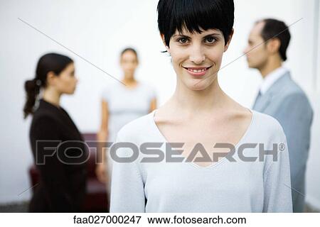 Young businesswoman smiling at camera, colleagues standing in background View Large Photo Image Stock Photo - Young businesswoman smiling at camera, colleagues standing in background. Fotosearch