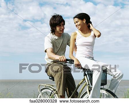 Young couple riding bicycle, female sitting on handlebars, looking over shoulder View Large Photo Image Stock Photo - Young couple riding bicycle, female sitting on handlebars, looking over shoulder. Fotosearch