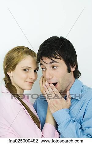Stock Image - Young couple together, woman looking at camera, man looking away and covering mouth with hand. Fotosearch