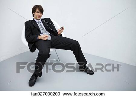 Young man in suit leaning back in chair, listening to mp3 player, smiling at camera, full length View Large Photo Image Stock Photo - Young man in suit leaning back in chair, listening to mp3 player, smiling at camera, full length. Fotosearch