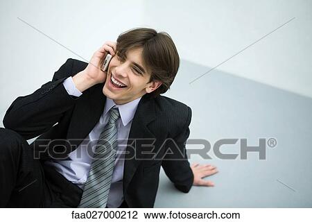 Young man in suit sitting on the ground, using cell phone, smiling View Large Photo Image Stock Image - Young man in suit sitting on the ground, using cell phone, smiling. Fotosearch