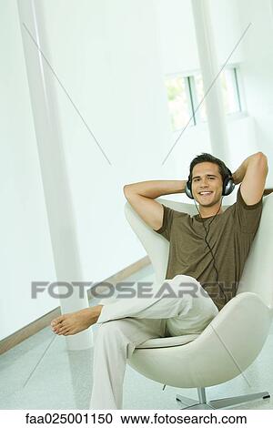 Young man listening to headphones with hands behind head, smiling at camera View Large Photo Image Stock Image - Young man listening to headphones with hands behind head, smiling at camera. Fotosearch