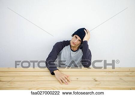 Stock Image - Young man sitting at table, listening to earphones, holding head, looking at camera. Fotosearch