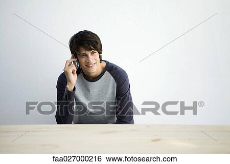 Young man sitting at table, using cell phone, smiling View Large Photo Image Stock Photograph - Young man sitting at table, using cell phone, smiling. Fotosearch