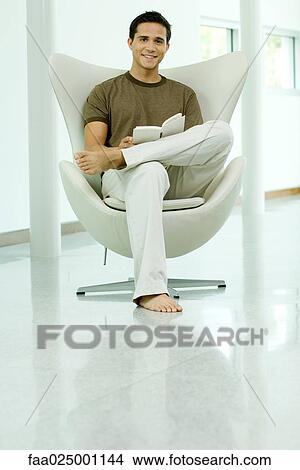 Young man sitting in chair holding book, smiling at camera, full length View Large Photo Image Picture - Young man sitting in chair holding book, smiling at camera, full length. Fotosearch