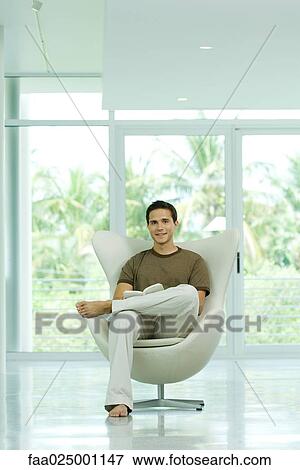 Young man sitting in chair holding book, smiling at camera View Large Photo Image Stock Photo - Young man sitting in chair holding book, smiling at camera. Fotosearch