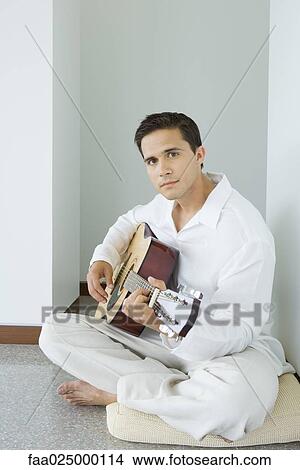 Young man sitting on cushion playing acoustic guitar, looking at camera View Large Photo Image Picture - Young man sitting on cushion playing acoustic guitar, looking at camera. Fotosearch