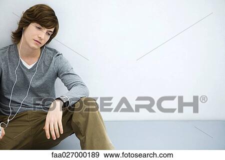 Young man sitting on the ground, listening to earphones, looking away View Large Photo Image Stock Photo - Young man sitting on the ground, listening to earphones, looking away. Fotosearch