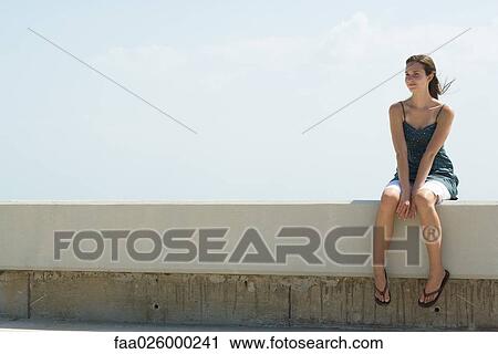 Stock Image - Young woman sitting on low wall outdoors, smiling, looking away. Fotosearch