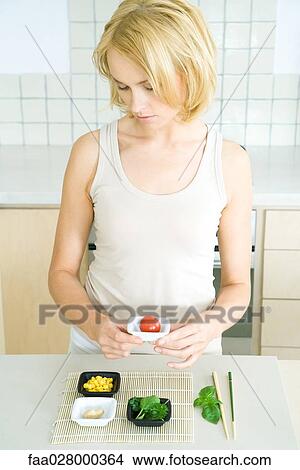 Young woman standing in kitchen, holding cherry tomato in small dish, looking down View Large Photo Image Picture - Young woman standing in kitchen, holding cherry tomato in small dish, looking down. Fotosearch
