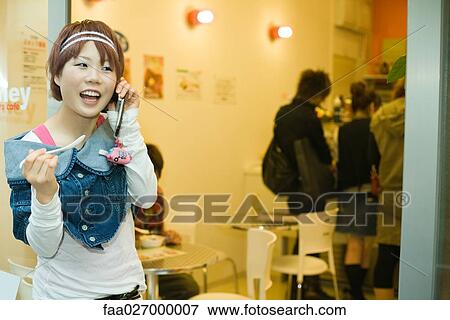 Young woman standing outside restaurant, using cell phone, holding spoon View Large Photo Image Stock Photo - Young woman standing outside restaurant, using cell phone, holding spoon. Fotosearch