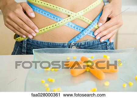 Young woman with measuring tapes wrapped around waist, plate of vegetables in foreground, cropped view View Large Photo Image Stock Image - Young woman with measuring tapes wrapped around waist, plate of vegetables in foreground, cropped view. Fotosearch