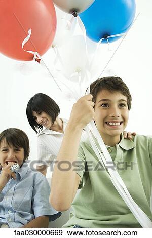 Boy holding helium balloons, smiling, mother and brother in background View Large Photo Image Stock Photo - Boy holding helium balloons, smiling, mother and brother in background. Fotosearch