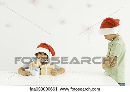 Stock Image - Boy holding stack of Christmas gifts, brother standing nearby, sulking, both wearing Santa hats. Fotosearch