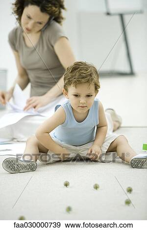 Boy playing with marbles while young mother uses phone and looks at paperwork in background View Large Photo Image Stock Photo - Boy playing with marbles while young mother uses phone and looks at paperwork in background. Fotosearch