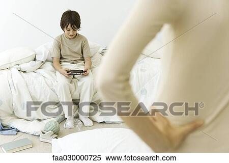Boy sitting in messy room, playing handheld video game, mother in foreground with hand on hip, cropped view View Large Photo Image Stock Photography - Boy sitting in messy room, playing handheld video game, mother in foreground with hand on hip, cropped view. Fotosearch