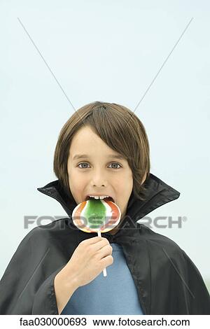 Stock Image - Boy wearing vampire cape, eating large lollipop, portrait. Fotosearch