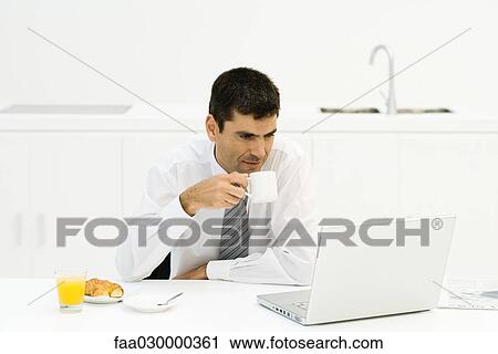 Businessman sitting at kitchen table, looking at laptop computer, drinking coffee View Large Photo Image Stock Image - Businessman sitting at kitchen table, looking at laptop computer, drinking coffee. Fotosearch
