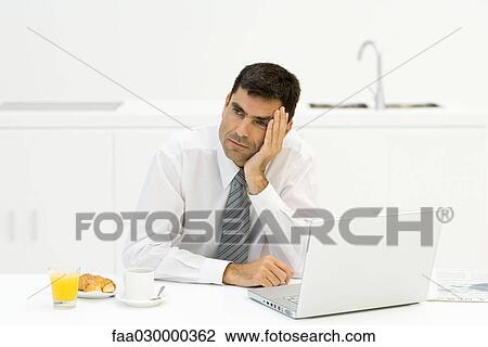 Stock Image - Businessman sitting in kitchen with breakfast and laptop, leaning head on hand, looking away. Fotosearch