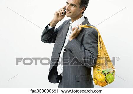 Stock Image - Businessman using cell phone, mesh bag of groceries slung over his shoulder. Fotosearch