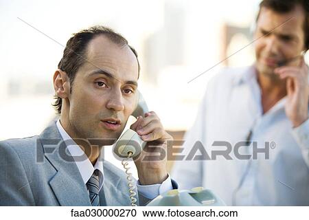 Stock Image - Businessman using landline phone, man in background using headset. Fotosearch