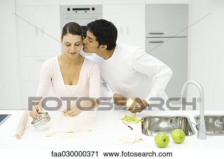 Stock Image - Couple cooking together, man kissing woman's cheek. Fotosearch