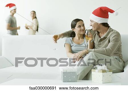 Couple sitting on sofa, drinking champagne, man wearing Santa hat View Large Photo Image Stock Image - Couple sitting on sofa, drinking champagne, man wearing Santa hat. Fotosearch