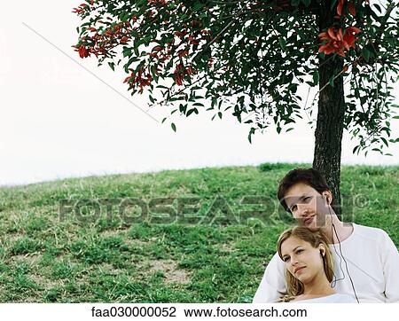 Stock Image - Couple sitting under tree, sharing earphones. Fotosearch